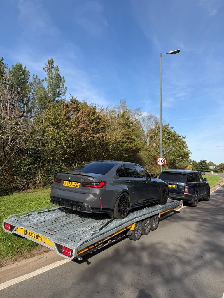 Dark gray BMW sedan loaded on a yellow and black car trailer on a paved road with trees and grass in background