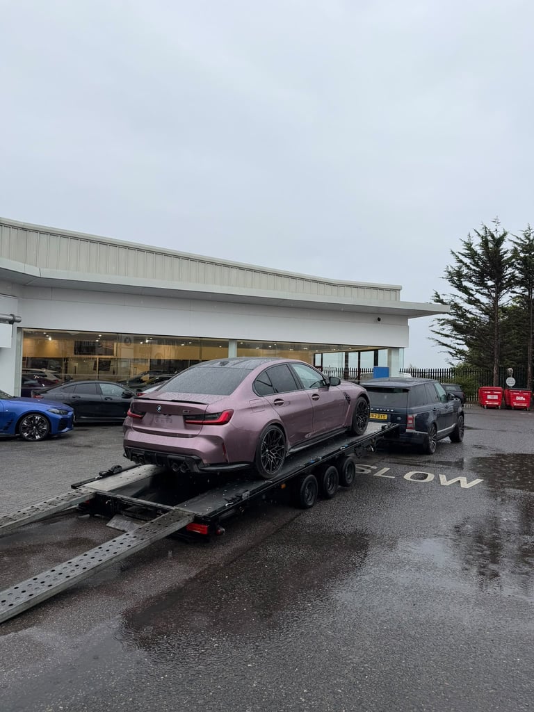 Pink BMW sedan being loaded onto a flatbed tow truck at an auto garage with other vehicles visible in the background