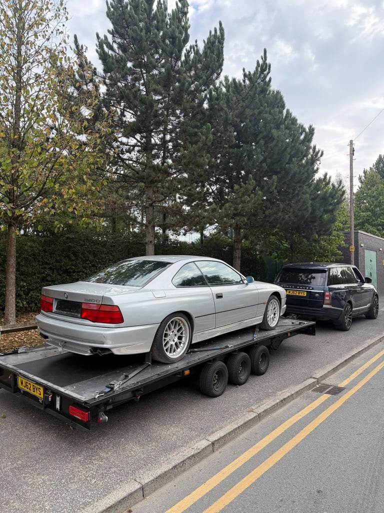 Silver BMW coupe being transported on a flatbed trailer on a residential street with trees in background