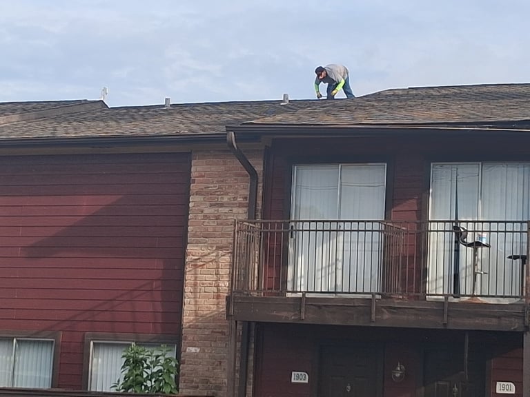 Roofer working on top of a red brick residential building with a balcony and multiple windows — apartment roof repair