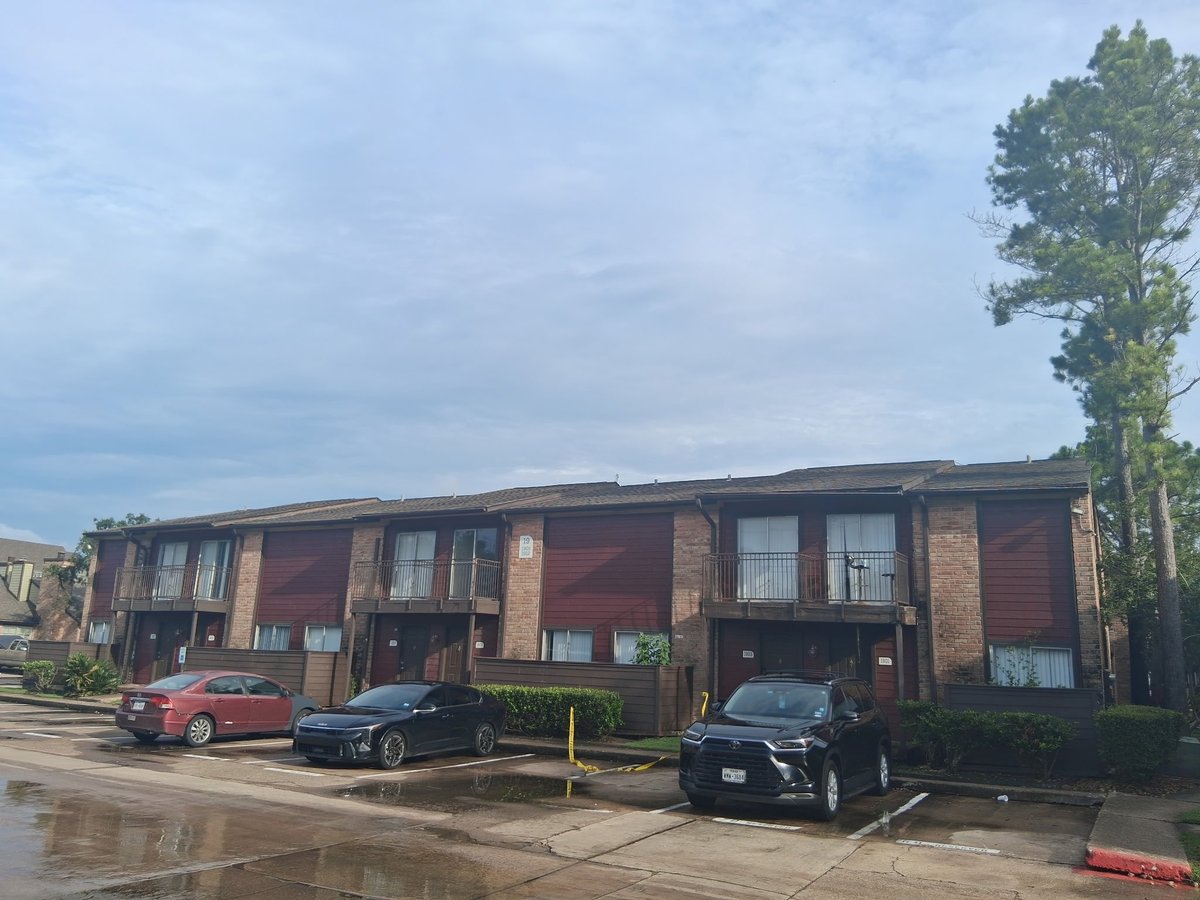 Red brick apartment building with cars parked in front and a tall tree on the right side — N1 apartment community