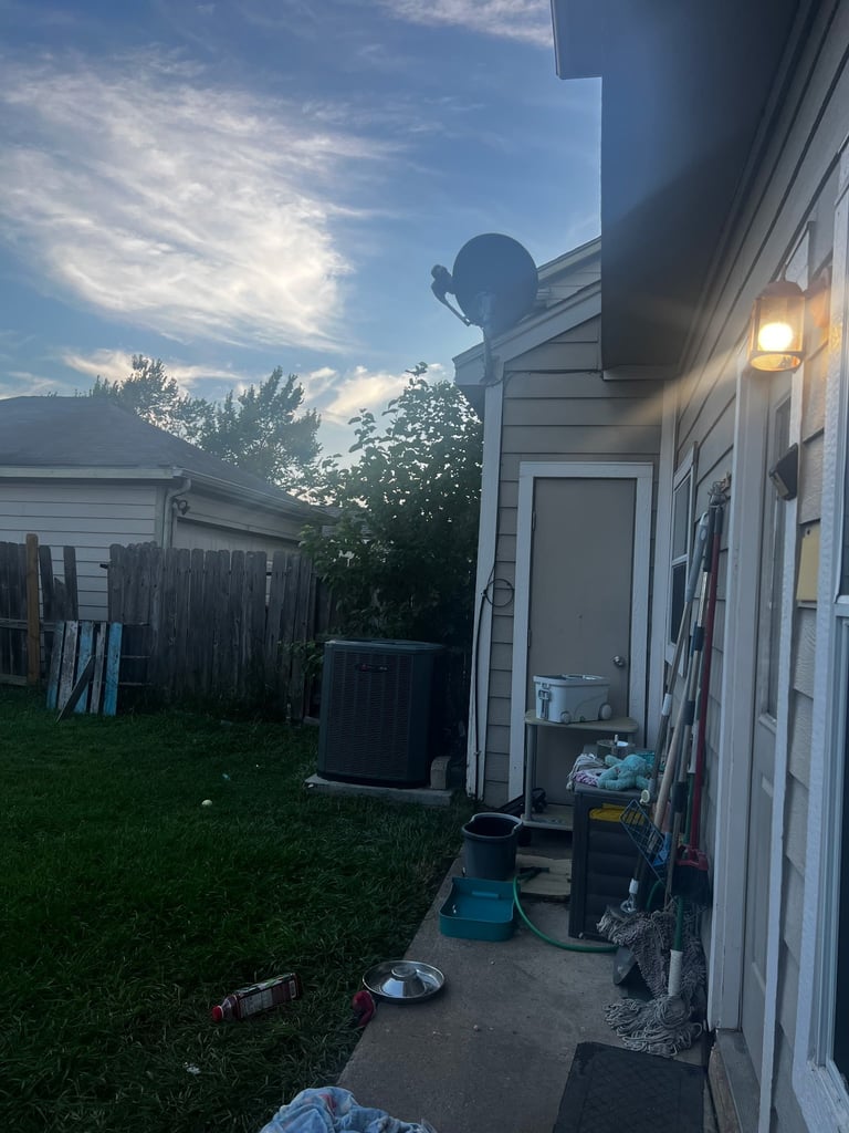 Backyard view of white house with satellite dish mounted on roof, green lawn, wooden fence, and storage containers at dusk
