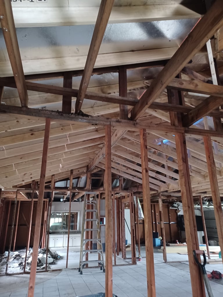 Interior framing of the home under reconstruction showing new wooden beams, metal roof panels, and an aluminum ladder on the concrete floor