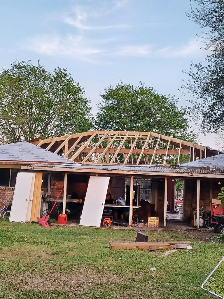 Mid-remodel: House roof stripped to exposed wooden frame and beams, with grass and trees surrounding the property