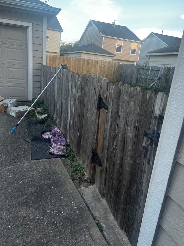 Weathered wooden fence alongside residential driveway with gray garage, suburban neighborhood homes in background