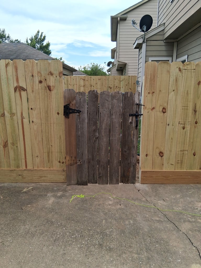 Wooden fence gate with weathered center panel flanked by two lighter-colored gate sections, with residential house and satellite dish in background