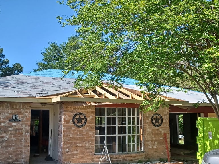 Mid-remodel: Red brick house with barred windows, Texas star decorations, and exposed roof framing under tall trees