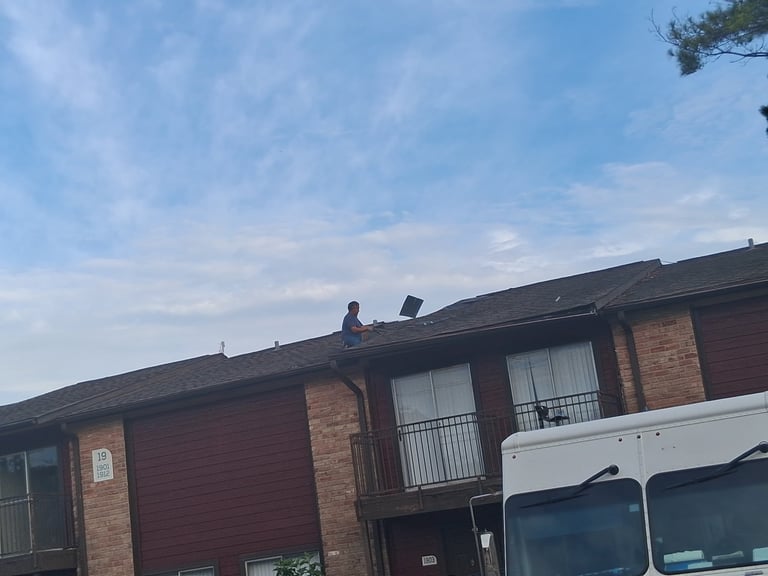 Person sitting on roof of brick residential house with vehicles parked in driveway below — apartment roof inspection