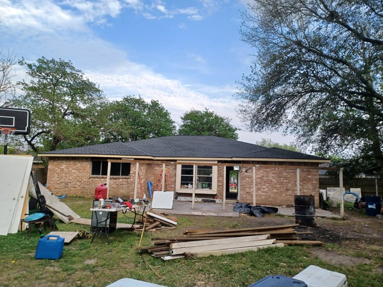 Back of the brick house mid-renovation with new roof, construction materials, tools, and wooden debris scattered in the yard