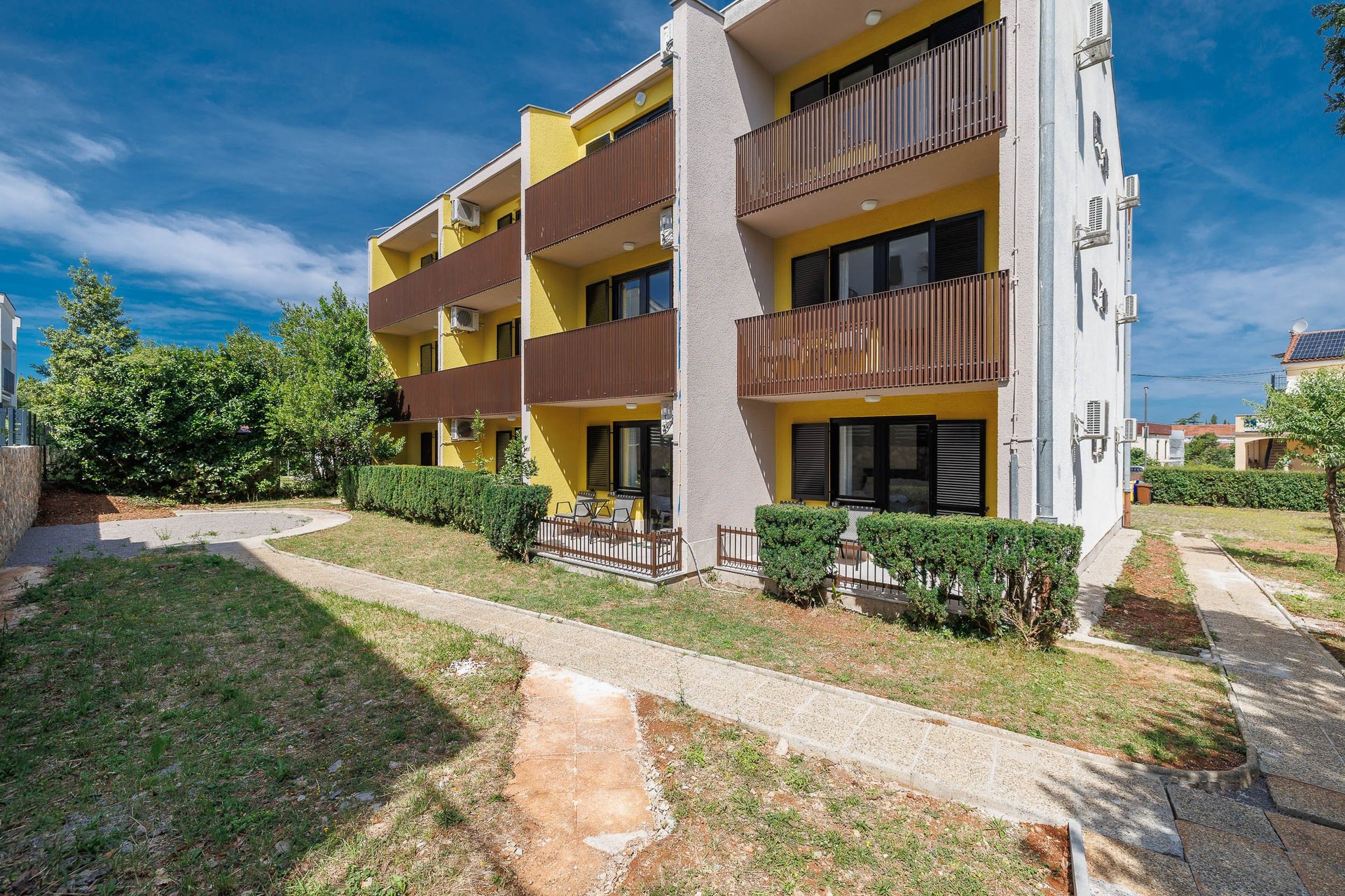 Modern apartment building with yellow and white facade, multiple balconies, landscaped front yard with lawn and shrubs under clear blue sky