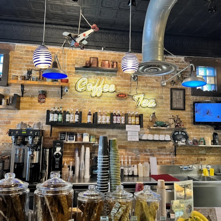 Interior of a coffee shop with brick walls, neon Coffee Tea sign, espresso machines, hanging pendant lights, and a person on an aerial rope above