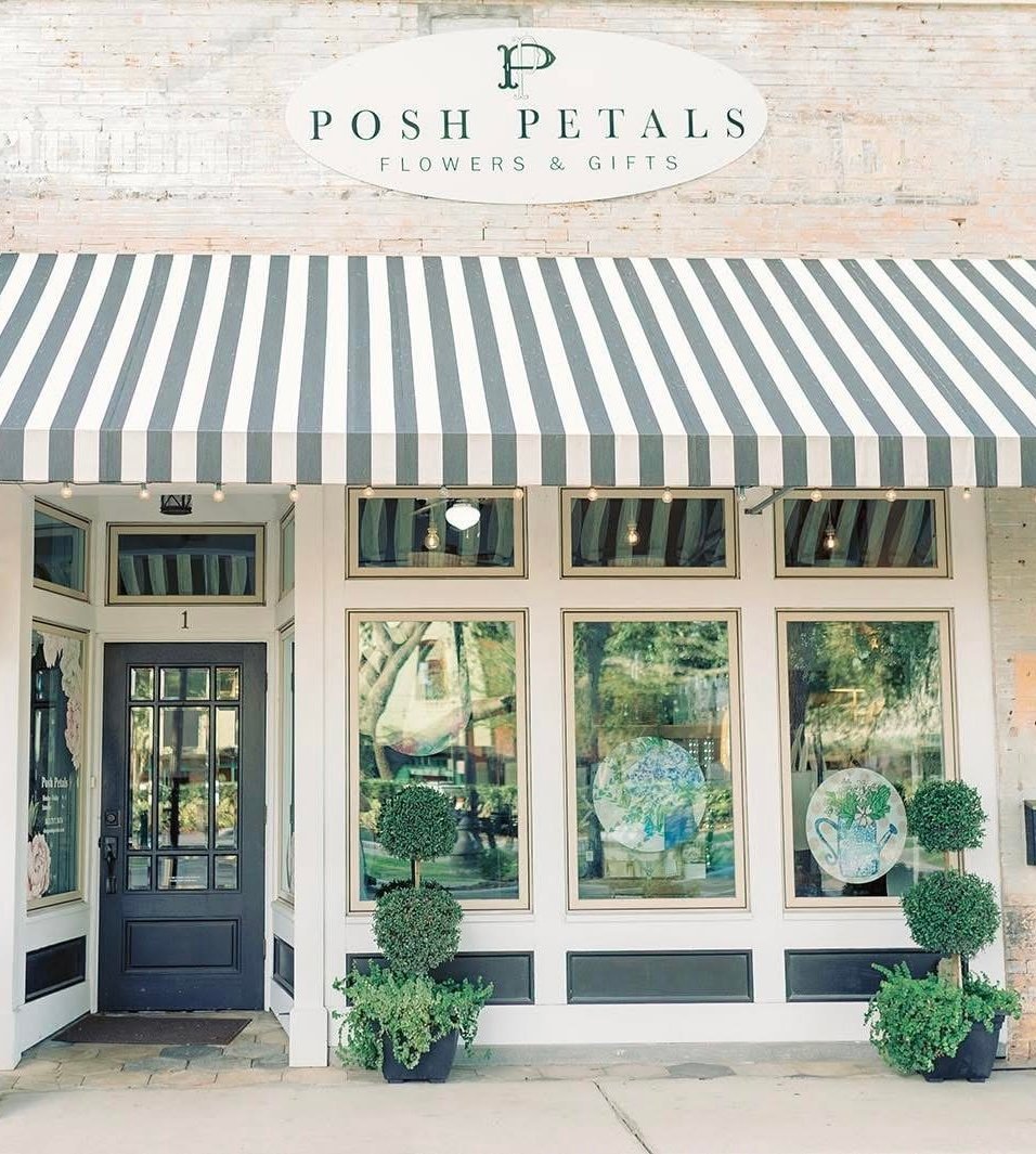 Posh Petals flower shop storefront with gray and white striped awning, large display windows, and green topiary plants by entrance