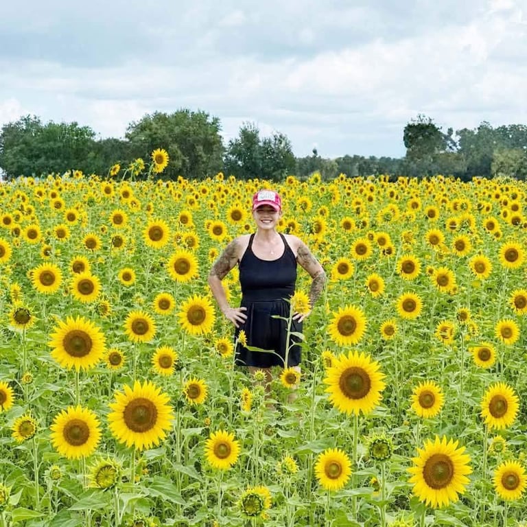 Woman in athletic wear standing in a vast sunflower field with green trees under a cloudy sky