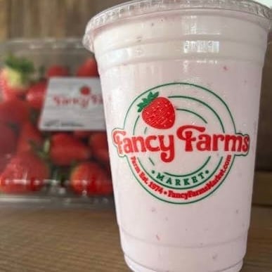Fancy Farms strawberry milk drink in a clear plastic cup with red strawberries visible in background