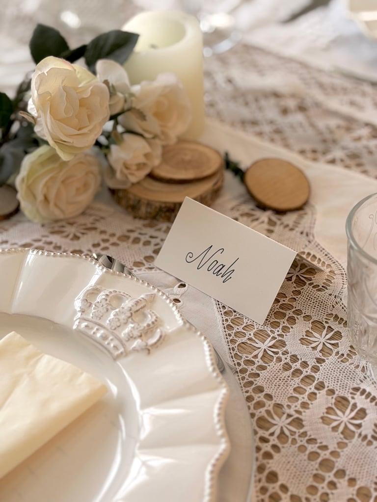 Elegant wedding table setting with cream roses, lace tablecloth, white plates, and a place card reading Noah