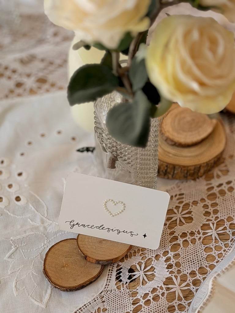 Elegant thank you card with dotted heart design on delicate lace tablecloth next to cream roses, wood slices, and burlap ribbon