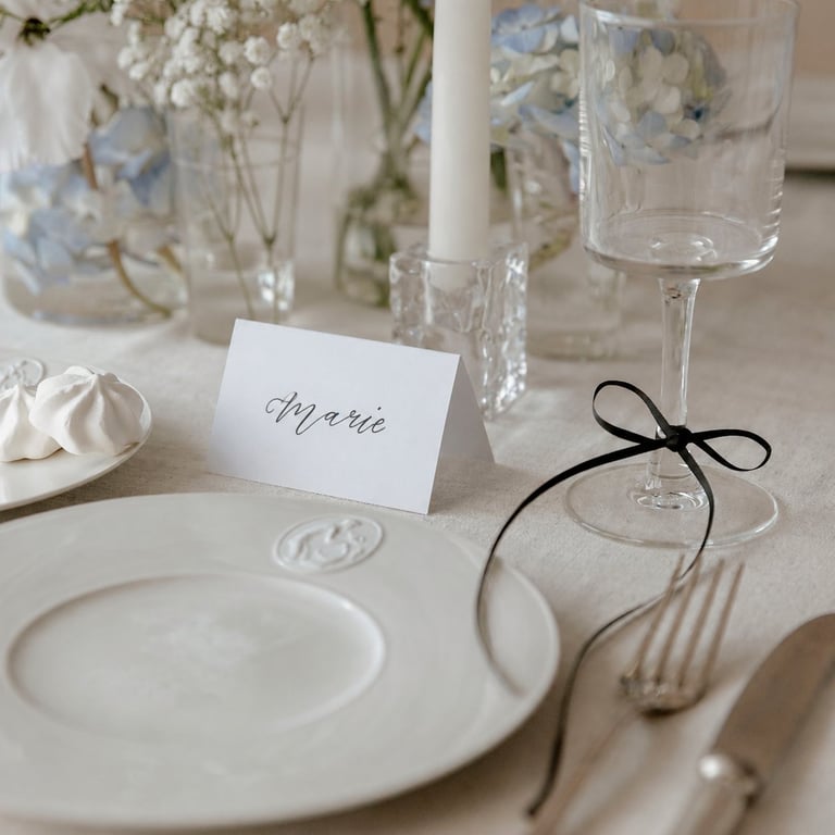 Elegant formal table setting with white plates, place card reading Marie, black ribbon napkin tie, crystal glassware, and white flowers