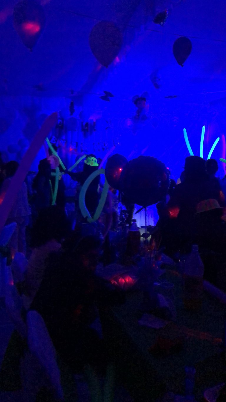 Crowd of people dancing at a neon ultraviolet party with glowing blue and pink lights, glow sticks, and balloons floating overhead