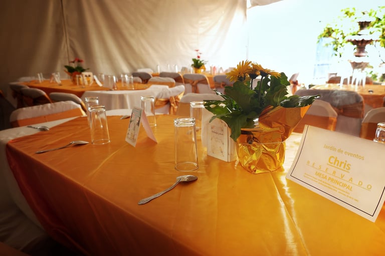 Elegant outdoor dining table with orange tablecloth, place settings, yellow flowers, and name cards under a tent