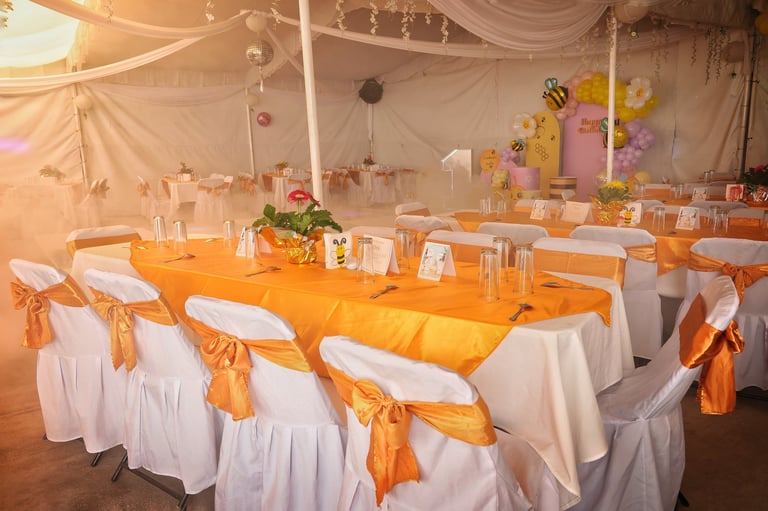 Elegantly decorated banquet hall with long tables draped in white linens and gold sashes, flower centerpieces, and hanging decorations for an event