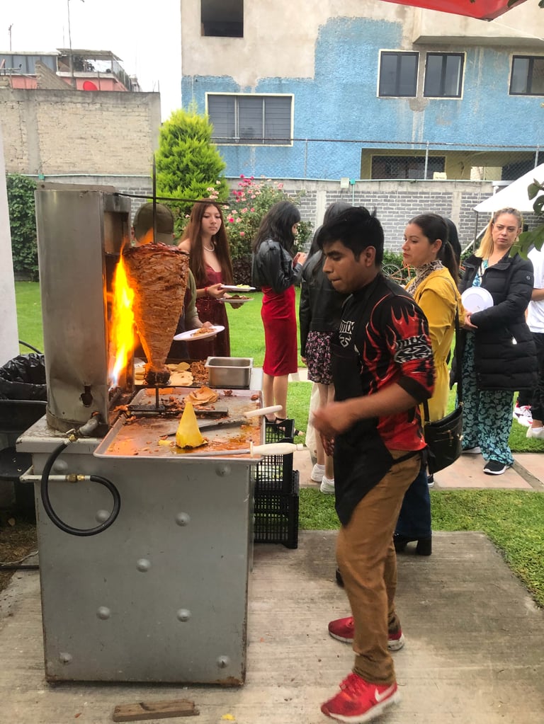 People gathered around a vertical rotisserie grill cooking meat in a residential backyard during an outdoor gathering