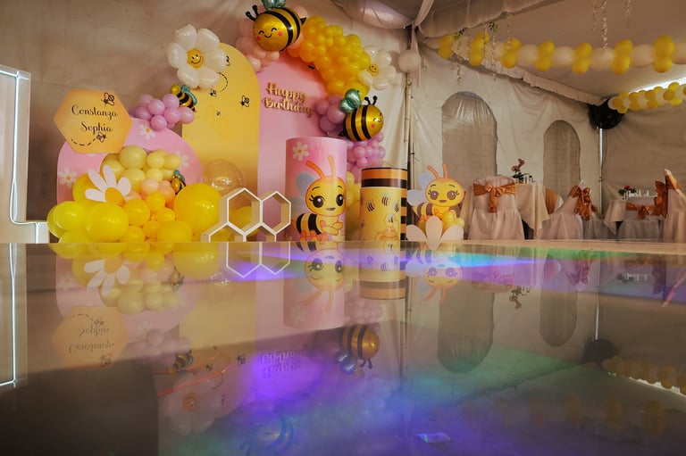 Colorful bee-themed party decoration setup with yellow and pink balloons, bee cutouts, and rainbow lighting reflecting on a polished floor