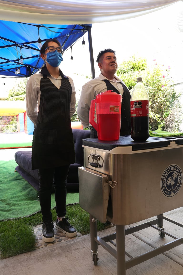 Two men in formal attire stand behind a stainless steel counter with red appliances under a blue shade canopy outdoors