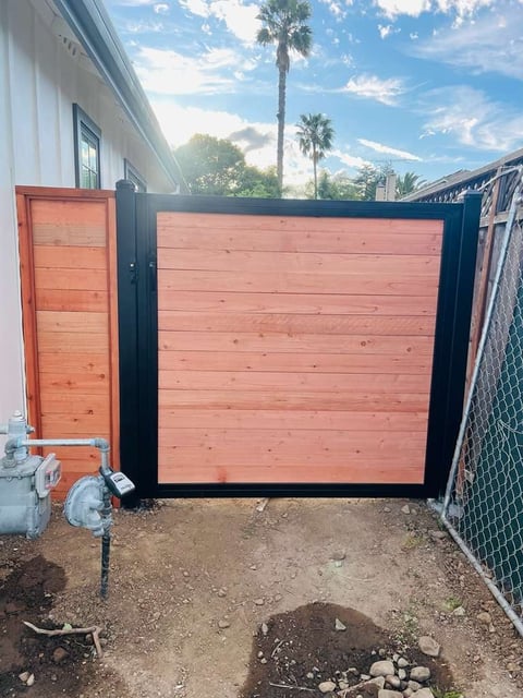 Modern wooden gate with black frame and red-cedar panels beside white building, tropical setting with palm trees visible