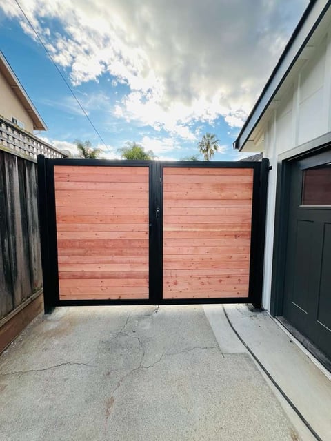 Modern driveway with double gate featuring red wood panels and black metal frames, leading to residential property under blue sky