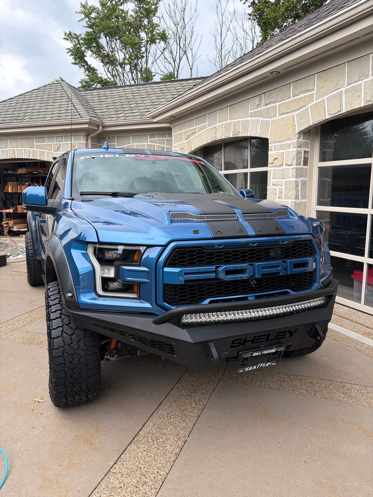 Blue Shelby Ford Raptor pickup truck with custom front bumper parked in driveway in front of stone house