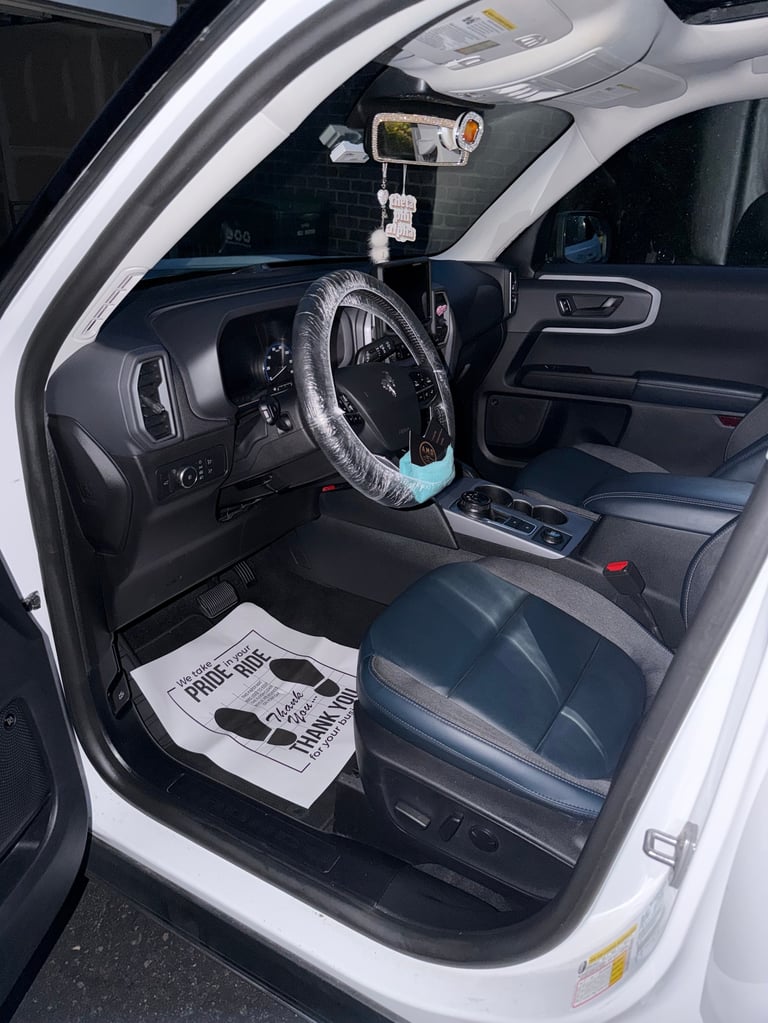 Interior view of white vehicle with open driver door, steering wheel with cover, dashboard, and protective plastic floor mat