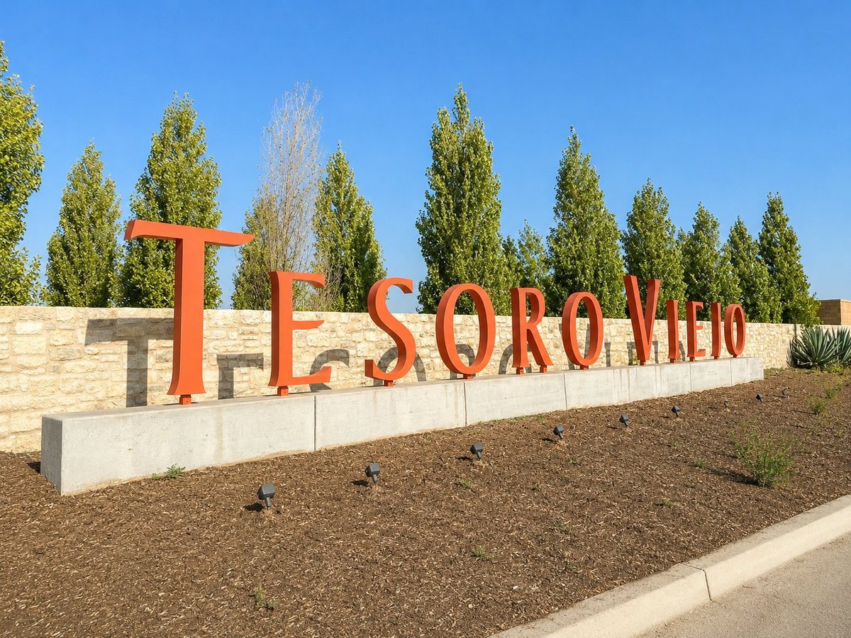 Large red metal TESORO VIEJO entrance sign in landscaped area with mature cypress trees
