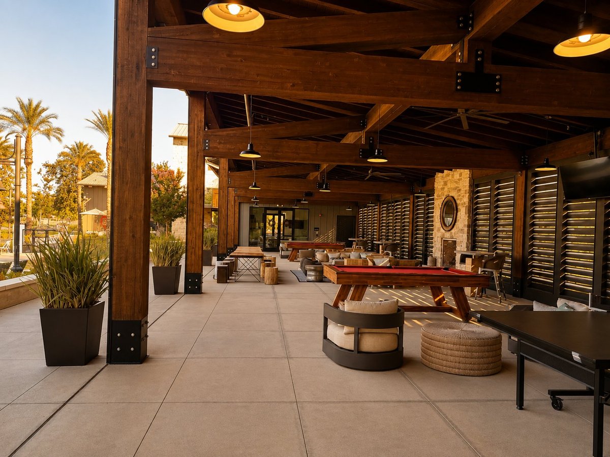 Modern covered outdoor patio with wooden beams, lounge seating, palm trees, and warm lighting at sunset