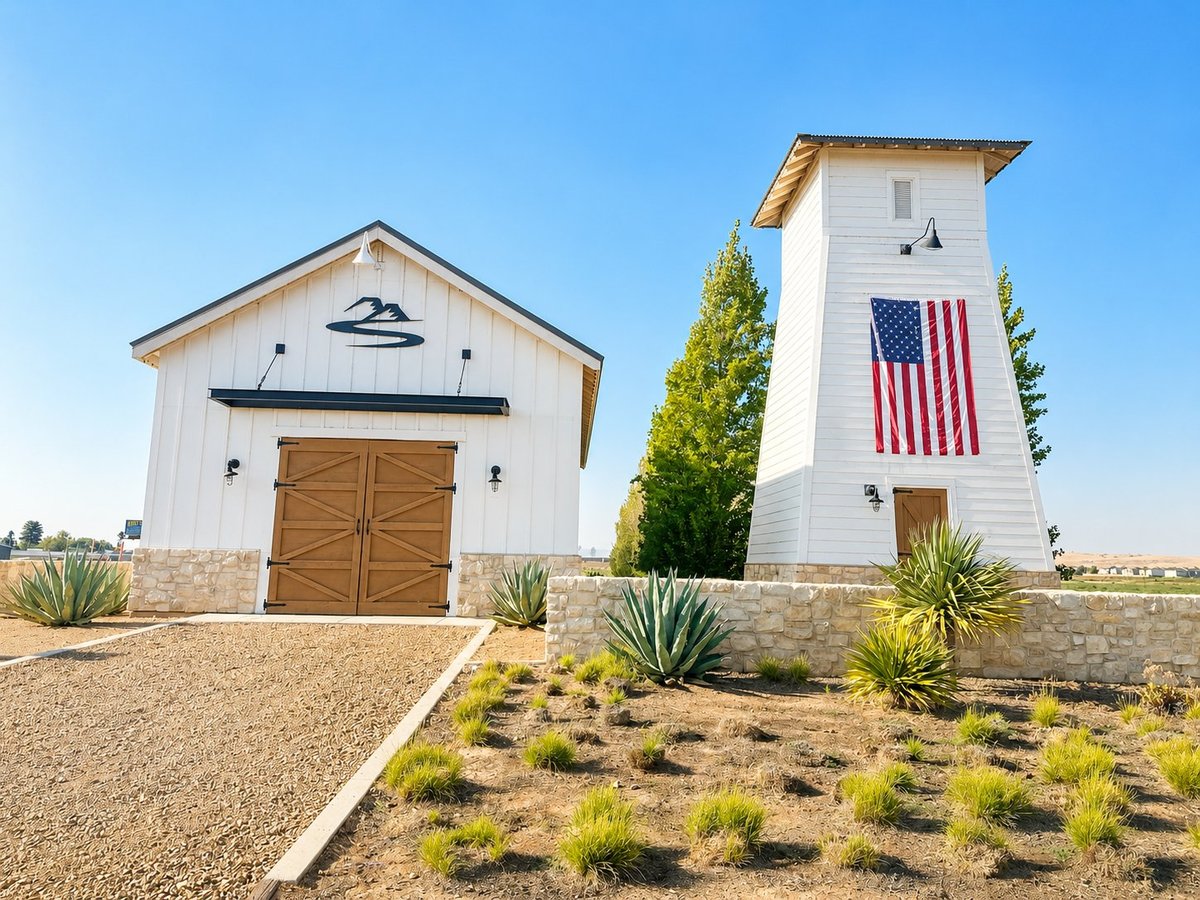 Two modern white buildings with stone bases in desert landscape — one barn-style structure with wooden doors and a tower building displaying American flag