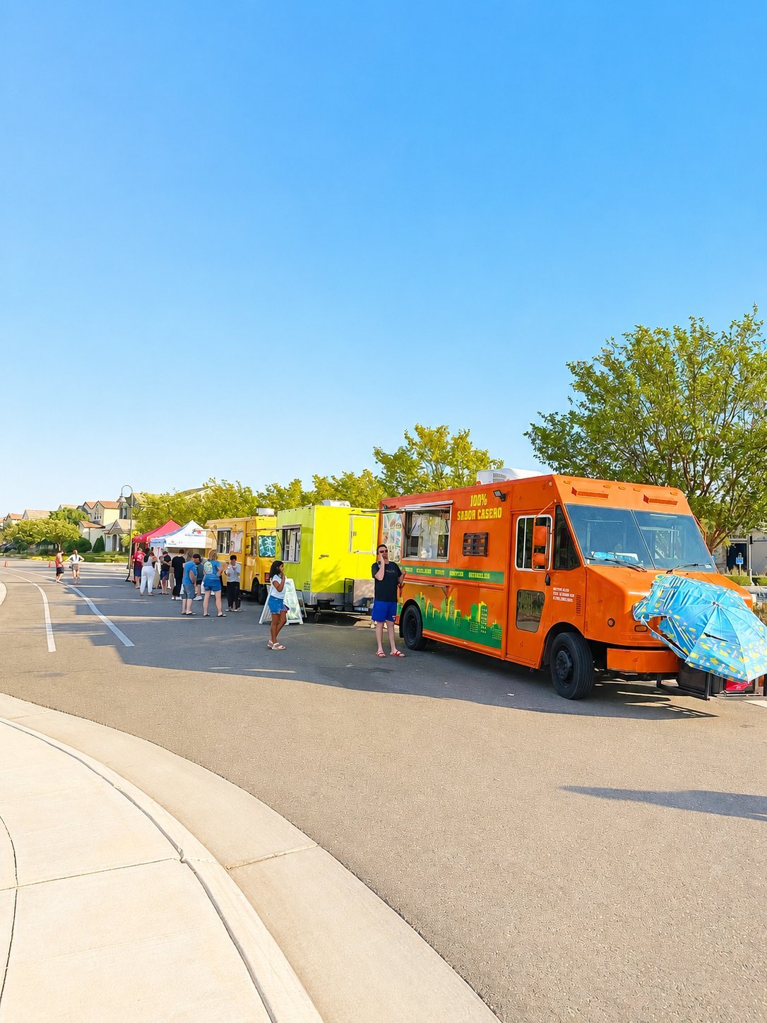 Food trucks lined up in a parking lot with customers waiting, featuring a bright orange and green truck in the foreground on a sunny day