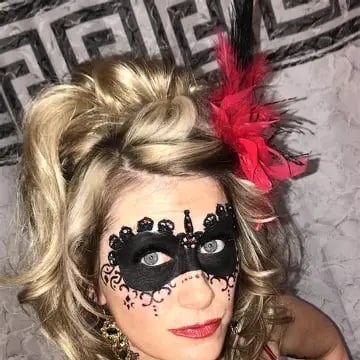 Woman wearing an ornate black lace masquerade mask with a red feather fascinator in blonde curled hair