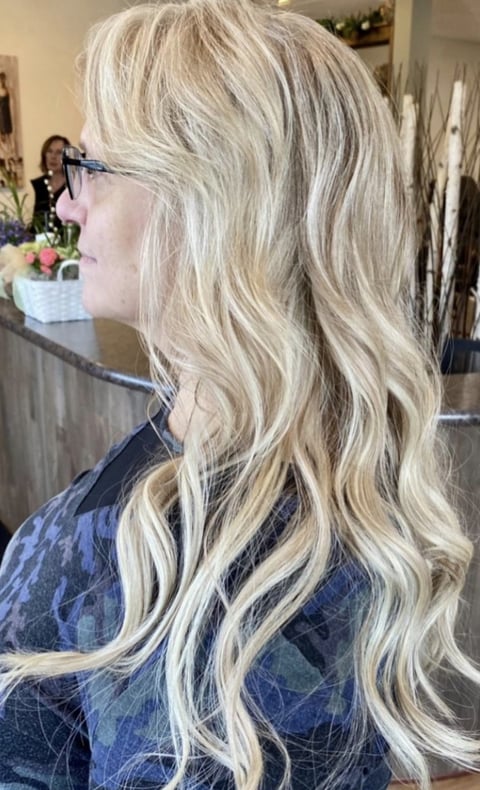 Woman with long blonde wavy hair, shown in profile view wearing glasses and a blue jacket in a salon setting