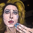 Portrait of a person with pale makeup and blonde hair, hand touching their face against a dark background