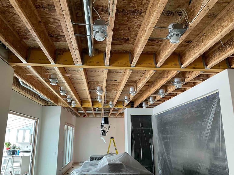 Interior renovation showing exposed wooden ceiling beams with recessed lighting fixtures installed in unfinished room