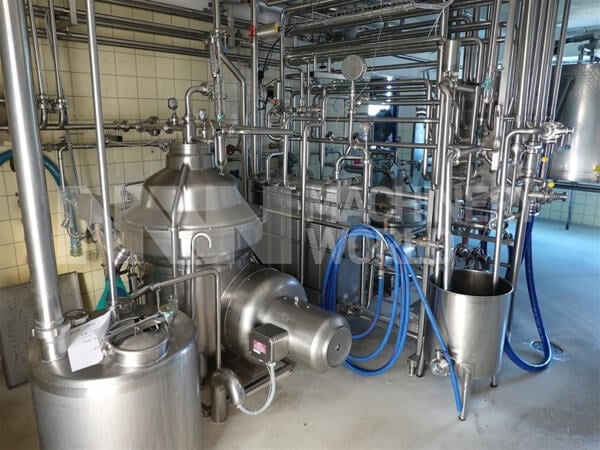 Industrial brewery or dairy processing facility with large stainless steel tanks, pipes, and blue hoses in a modern production area