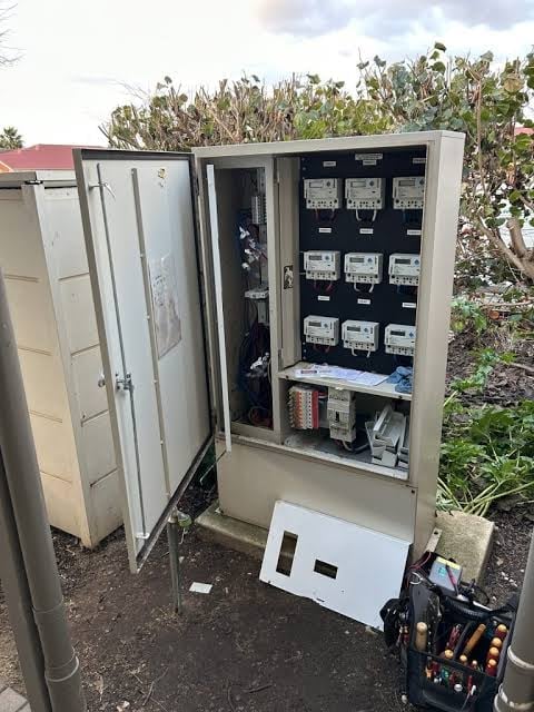 Open electrical meter panel cabinet mounted outdoors with multiple utility meters, wiring, and equipment visible inside