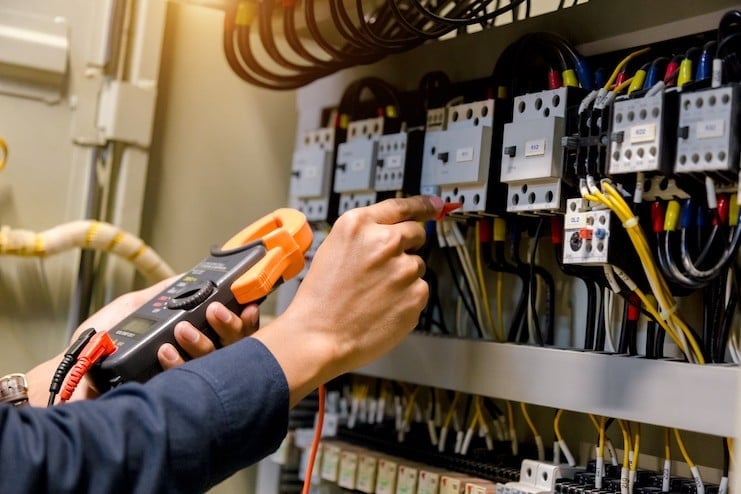 Electrician holding digital multimeter while testing electrical switchboard with circuit breakers and wiring
