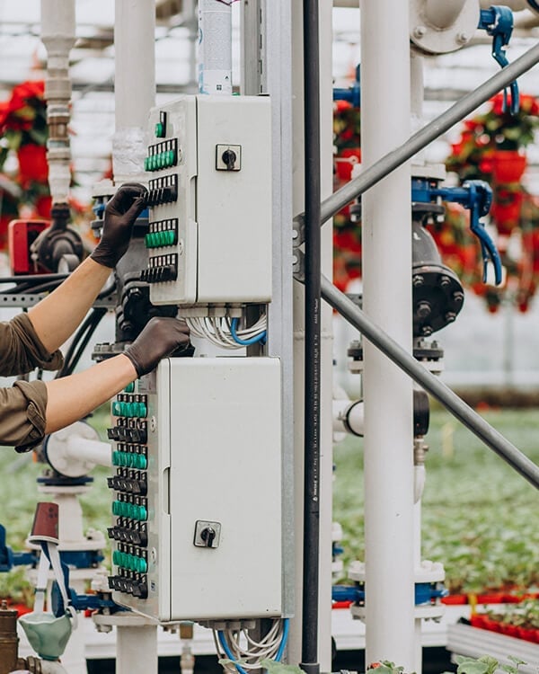 Technician wearing black gloves installing electrical panel on industrial structure with red flowers and equipment in background
