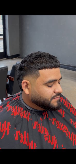 Man with short dark hair and beard wearing a red and black patterned shirt in a barber shop