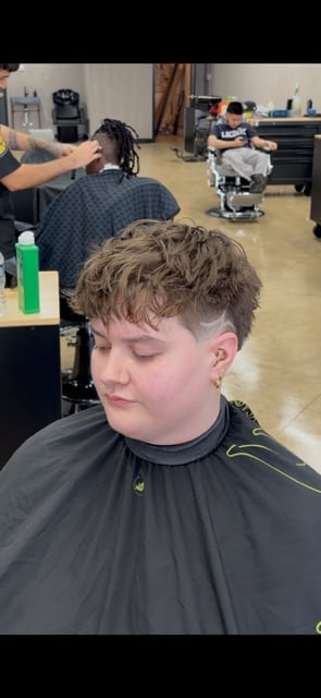 Young person getting a short textured haircut at a barbershop, wearing a black cape, with barber visible in background