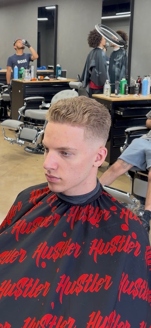 Young man with fresh fade haircut wearing red and black Hustler shirt in barbershop with mirrors and styling stations