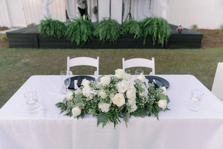 Outdoor wedding reception table with white tablecloth, white flowers and greenery centerpiece, white chairs, and green planter boxes in background