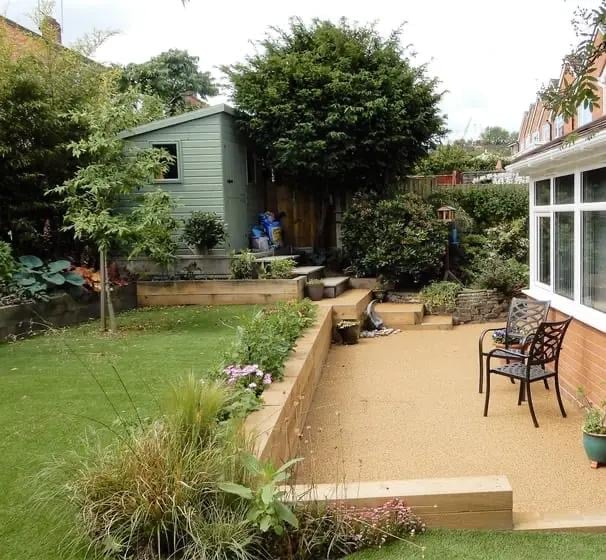 Backyard garden with gravel patio, ornate metal chair, raised beds, small garden shed, and lush green lawn surrounded by mature trees and hedges