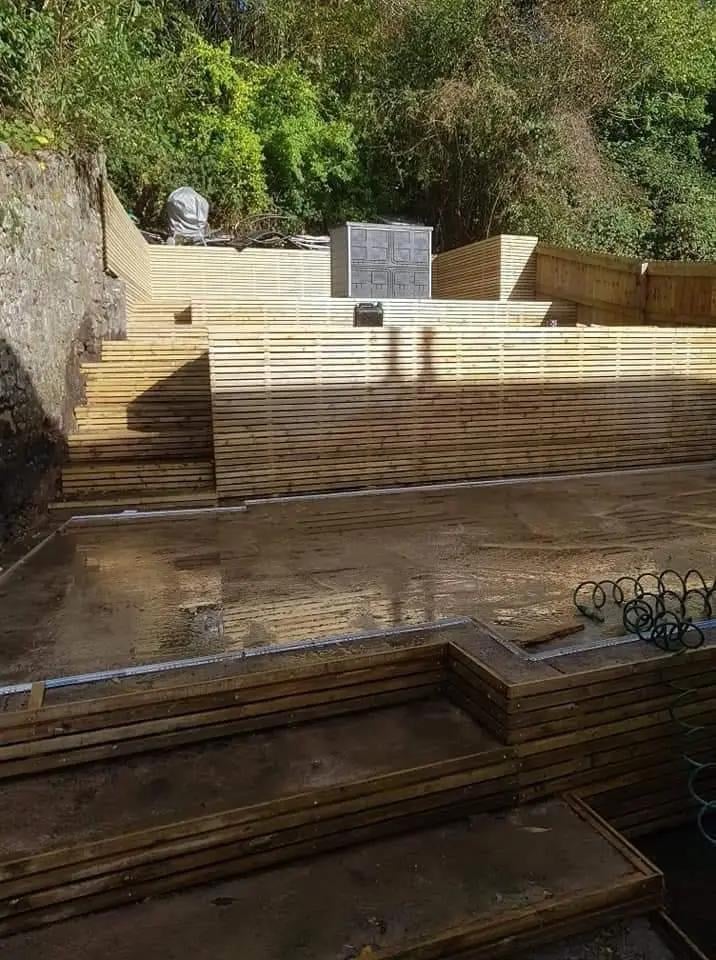 Wooden deck under construction with corrugated metal wall backdrop and overgrown vegetation, surrounded by building materials and bicycle racks