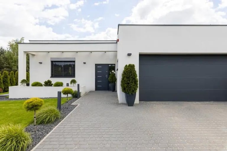 Modern white house with dark gray garage door, paved driveway, manicured lawn, and landscaping against blue sky